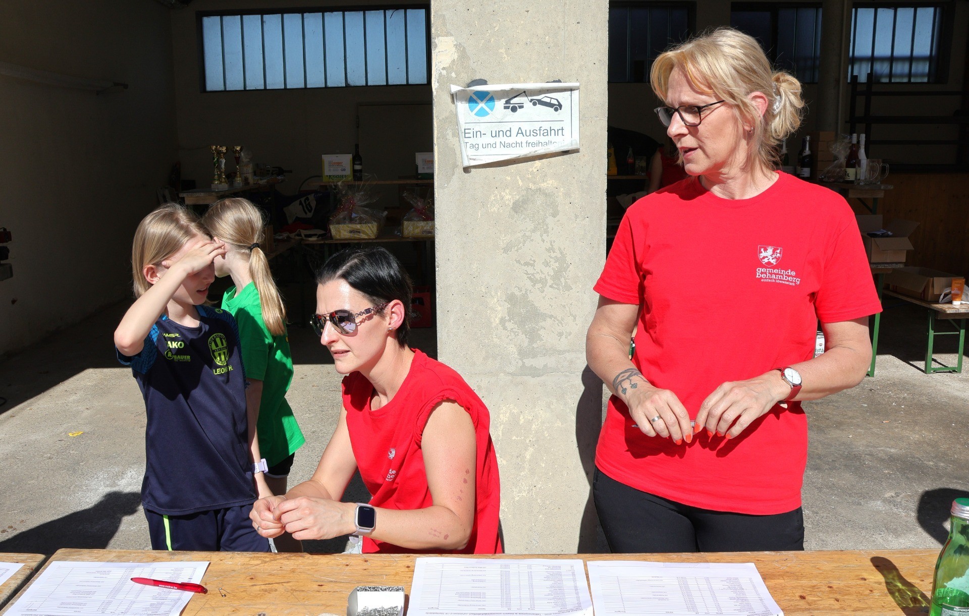 Eine Frau in Rot sitzt an einem Schreibtisch. Im Hintergrund ist eine andere Person zu sehen, die ebenfalls an einem Schreibtisch sitzt.