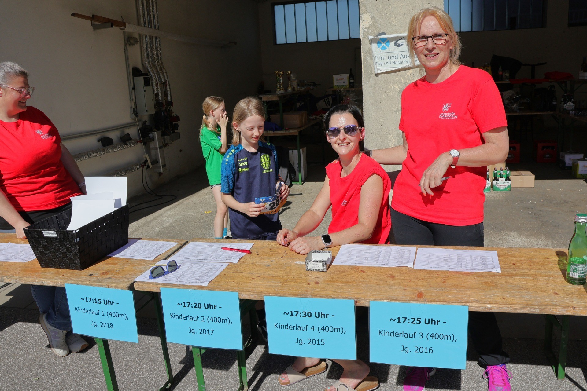 Eine junge Sportlerin mit Rot-Blau-Trikot sitzt an einer Tischplatte mit Zeitangaben für einen Wettlauf. Getränke stehen daneben.