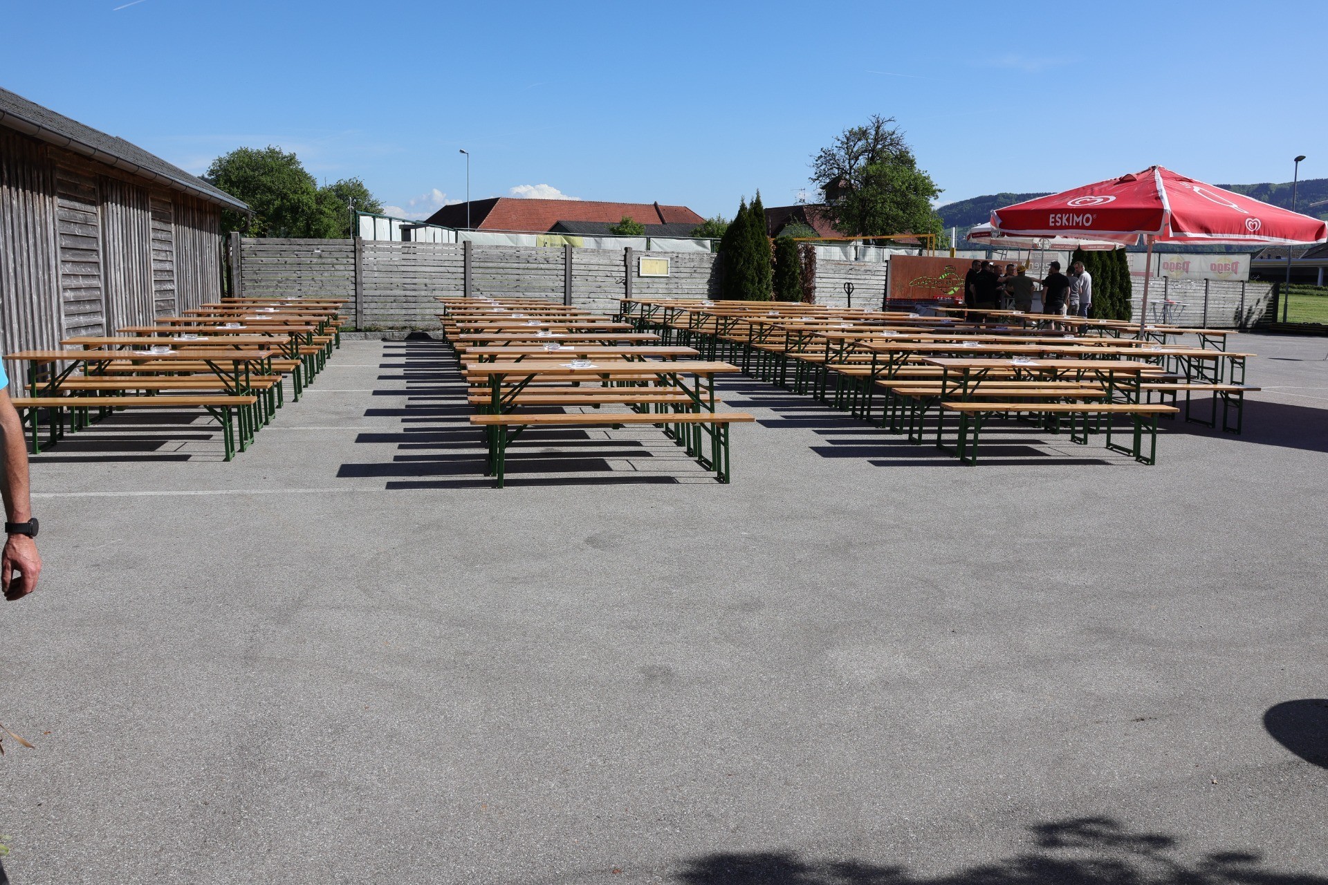 Flatternde Tische und Bänke auf einer Pflasterfläche vor einem Biergarten. Ein Getränkewagen steht im Hintergrund.