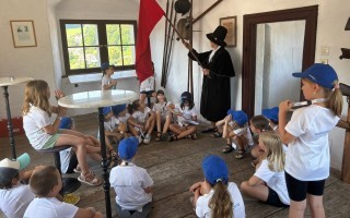 Kinder sitzen in einem Raum um einen Tisch und einen Mann in Schwarz mit Habicht.