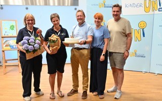 Ein Paar Menschen hält eine blaue Vase mit Blumen. Im Hintergrund ein grauer Raum.