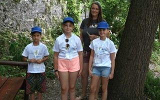 Vier Kinder in T-Shirts und Shorts stehen in einem Garten. Sie tragen blaue Mützen und Tragen jeweils eine grüne Tasche.