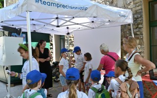 Kinder schauen unter einem Sonnensegel auf einen mobilen Ständer, vermutlich bei einer Veranstaltung.