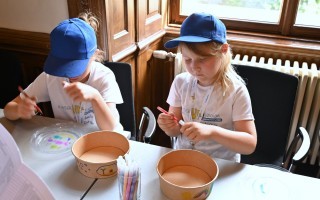 Zwei Kinder sitzen an einem Tisch und gestalten mit Buntstiften Pappschalen.