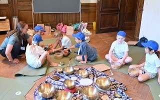 Kinder sitzen auf einem Teppich und üben mit Klangschalen. Eine erwachsene Person begleitet sie.