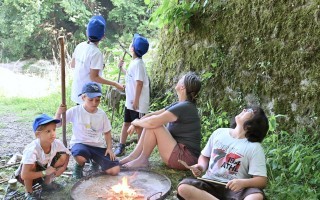 Kinder sitzen um einen Feuerkorb im Wald. Ein Junge versucht, ein Feuer zu entfachen, andere beobachten.