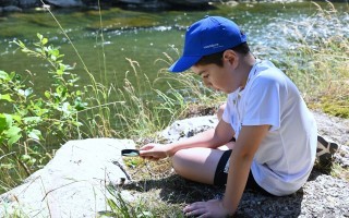 Ein Junge sitzt an einem Felsvorsprung und betrachtet mit einer Lupe eine Pflanze.
