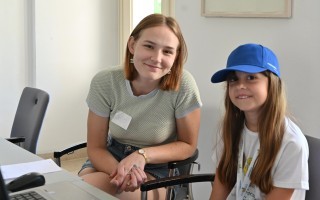 Lächzende junge Frau mit braunen Haaren sitzt in einem Büro. Sie trägt ein hellgraues T-Shirt, Jeans und einen blauen Baseballcap. Vor ihr steht ein Schreibtisch mit Papieren.