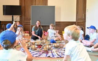 Kinder in einer Yogastunde mit Klangschalen. Eine Frau leitet die Gruppe in einem Raum mit einer Whiteboard-Leinwand.