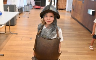 Ein Kind trägt ein glänzendes, rüstiges Brustpanzer-Kostüm in einem Flur mit Holzfußboden.