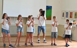 Mehrere Kinder und ein Erwachsener benutzen Smartphones auf einem hellen Holzboden. Im Hintergrund hängt ein farbenfrohes Kunstwerk an der Wand.