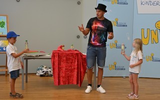 Drei Kinder stehen in einem Raum mit einer Staffelei. Ein Kind zeigt mit einer Flasche Wasser den anderen.