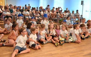Sitzende Kinder und Erwachsene in einem Raum. Einige tragen blaue Mützen, viele Kinder in hellen Kleidungsstücken sitzen auf dem Boden.