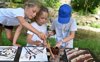 Drei Kinder malen mit Farben auf weiße Tafeln im Freien. Sie tragen T-Shirts.