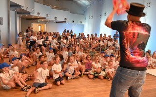 Kinder sitzen in einem Raum und schauen zu einem Mann mit einem Feuer-T-Shirt. Der Raum hat hohe Decken und viele Fenster.