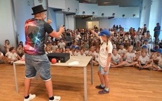 Ein Mann erklärt einer Kindesfigur auf einem Tisch etwas. Im Hintergrund sitzt eine Gruppe von Kindern auf dem Boden und hört zu.