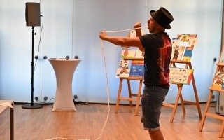 Mann zieht weiße Seilspanne durch Raum, mehrere Staffeleien mit bunten Gemälden im Hintergrund.