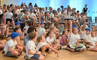Kinder sitzen auf einem Boden in einem Raum, einige tragen blaue Mützen. Es ist eine Gruppe von Kindern.