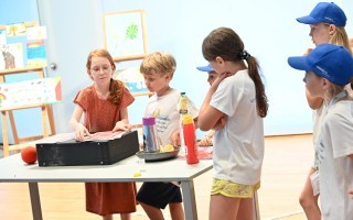 Mehrere Kinder untersuchen einen Tisch mit zwei Plastikflaschen und einem Blatt Papier in einem Klassenzimmer.