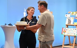 Zwei Personen stehen bei einer Veranstaltung. Eine Person hält eine Geburtstagstorte in der Hand. Im Hintergrund steht ein Kinderbauwagen und einige farbige Zeichnungen.