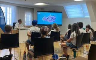 Schüler sitzen an einem Tisch in einem Klassenraum. Ein Mann steht am Whiteboard.