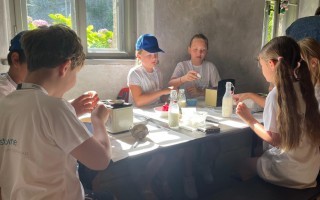 Mehrere Kinder sitzen an einem Tisch und essen oder trinken. Es ist ein sonniger Tag.