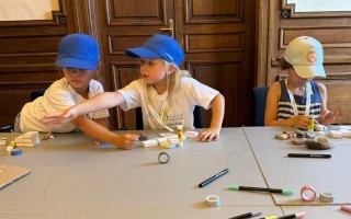 Drei Kinder bauen mit Holzstämmen an einem Tisch. Sie tragen T-Shirts und halten Schreibgeräte bereit.