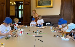 Kinder basteln an einem Tisch mit Papier, Stiften und kleinen Figuren in einem Raum mit einem Fenster.