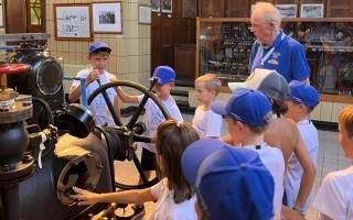 Kinder schauen einem älteren Mann beim Betrachten einer alten Maschine zu. Der Raum ist mit Technik gespickt.