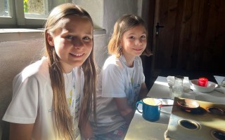 Zwei Mädchen mit langen Haaren sitzen an einem Tisch. Sie tragen T-Shirts und blicken in die Kamera. Auf dem Tisch stehen ein Becher, Besteck und Kerzen.