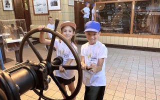 Drei Kinder in weißen T-Shirts stehen vor einem alten, schwarzen Bedienpult mit einem großen, rot lackierten Handrad.