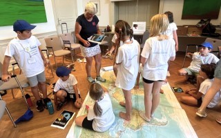 Kinder sind um eine Karte herum gruppiert, eine Frau sitzt daneben. Ein blaues T-Shirt und eine blaue Mütze sind zu sehen.