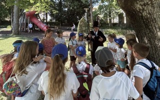 Kinder in blau, um einen Mann mit Tamburin herum, vor einem Baum.