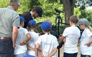 Ein Mann zeigt einer Gruppe von Kindern etwas, die in weißen T-Shirts mit dem Aufdruck 