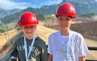 Zwei Kinder mit Bauarbeiterhelmen, die vor einem Zaun stehen. Sie tragen T-Shirts mit dem Aufdruck 