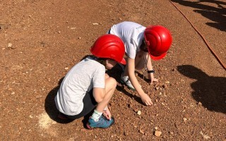 Zwei Kinder in Schutzhelmen graben mit Schaufeln in der Erde.