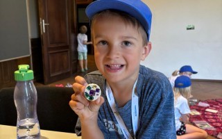 Ein Junge lächelt und hält ein buntes Glasobjekt in der Hand. Im Hintergrund sitzen weitere Kinder.