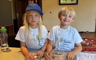 Zwei Kinder in weißen T-Shirts sitzen an einem Tisch und spielen mit kleinen bunten Perlen in einer Schale.