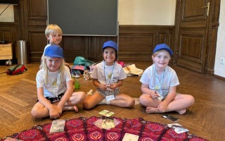 Vier Kinder sitzen auf einem Teppich und spielen mit Karten. Sie tragen helle Kleidung und Rucksäcke.