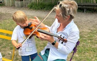 Ein Junge spielt eine Geige, ein Mann lehrt ihn, sitzt draußen.