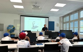 Schulklasse mit Kindern am Computer. Ein Lehrer steht vor einem Bildschirm mit einer 3D-Darstellung eines Pfeils.