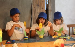 Drei Kinder bauen mit Papier und Stiften einen gelben Vogel zusammen an einem Tisch.