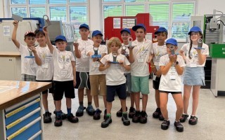 Schulkinder mit Schraubenschlüsseln vor einem Schreibtisch in einer Werkstatt. Einige tragen blaue Overalls, andere sind in hellen Farben gekleidet.