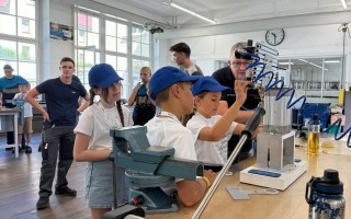 Schüler beobachten einen Tischdrucker in einem Klassenzimmer mit dunklem Boden. Eine Feder wird im Hintergrund gezeigt.