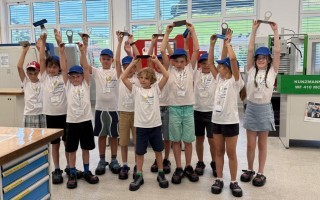 Kinder in einem Klassenzimmer, einige mit blauen Regenmänteln, stehen in einer Reihe. Im Hintergrund ist eine weiße Wand mit verschiedenen Bügeln und Papierordnern zu sehen.