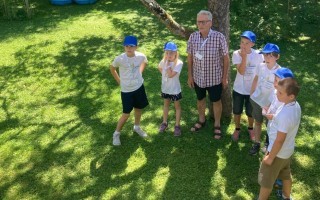 Mehrere Kinder in T-Shirts und Shorts stehen im Grünen zusammen. Ein Mann mit Brille ist ebenfalls dabei. Schatten fallen auf den Rasen.
