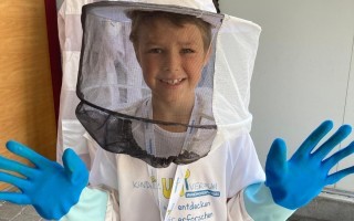 Junge mit Schutzanzug und Handschuhen vor einem Bienenstock.