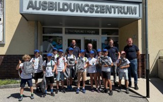 Eine Gruppe von Kindern und einem Erwachsenen lächeln auf einem Bild vor einem Gebäude. Sie tragen blaue Mützen und haben die Aufschrift 