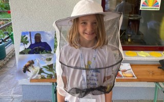 Junge Frau mit Bienenhelm vor einem Informationsstand mit Bienenbildern.