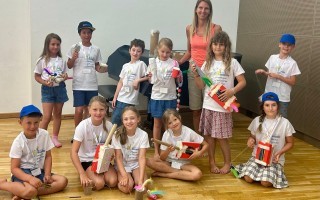 Sieben Kinder und eine Frau sitzen auf dem Boden in einem Raum. Die Kinder tragen blaue Mützen und T-Shirts. Sie halten Geschenke mit Schleifen in den Händen.